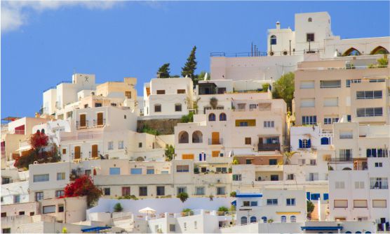 Vista de edificios y casas blancas con muchas ventanas acristaladas y con persianas azul marino, amarillo y tonos naranjas. Todas ellas tienen terrazas en sus azoteas. Entre ellas se ven árboles, plantas y enredaderas con flores de color fucsia. Los edificios están sobre  una colina. La imagen ha sido tomada desde abajo en un día soleado, contrastando  un cielo azul celeste con el blanco de las casas y una nube blanca.
